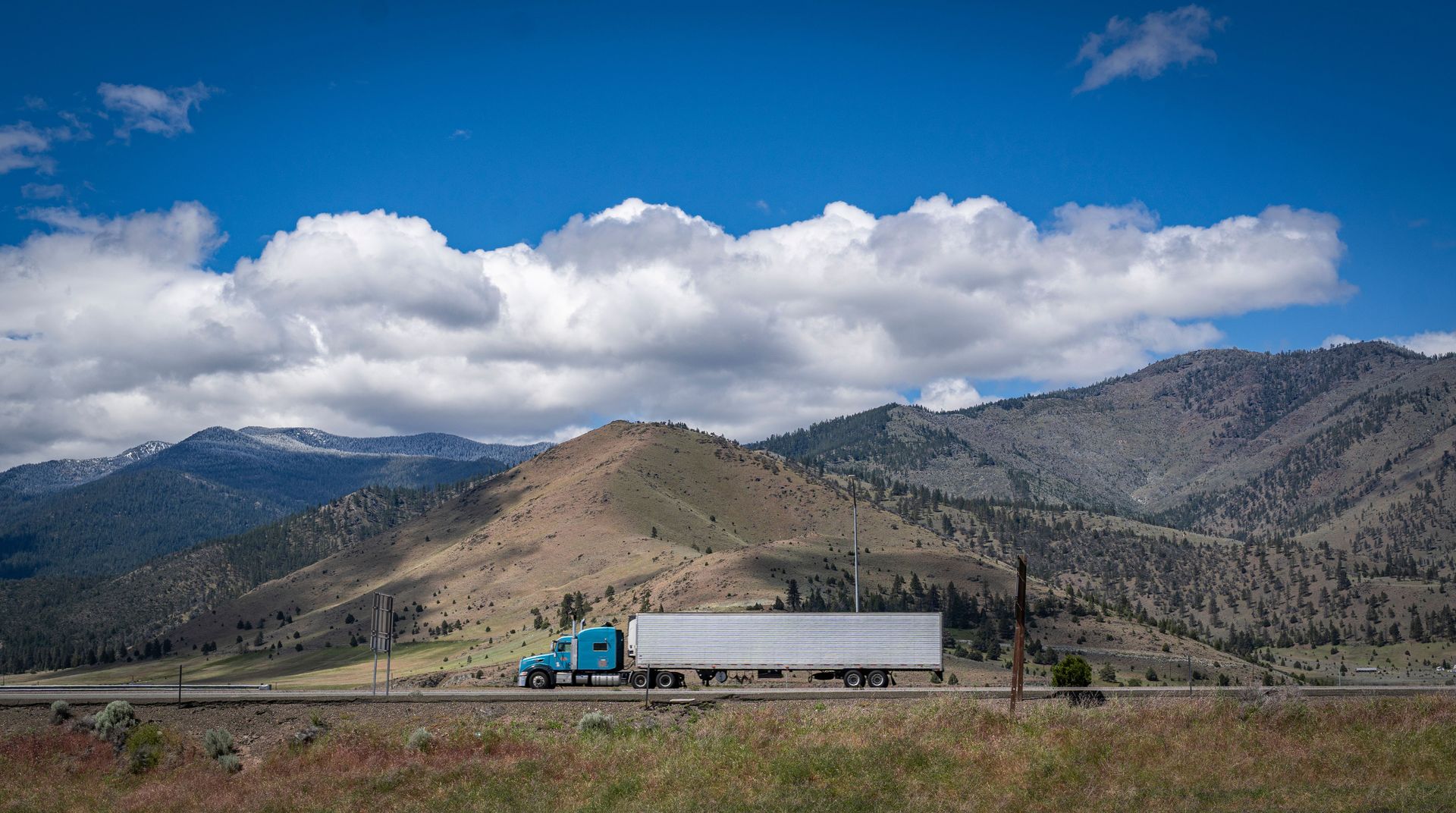 A blue semi-truck travels along a scenic highway with mountains and cloudy skies in the background.
